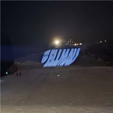 Eine helle Projektion des Wortes "Ellen" auf einer verschneiten Piste bei Nacht. Im Hintergrund sind Lichter und ein schneebedeckter Hang sichtbar.