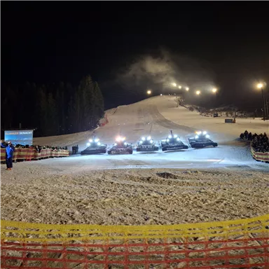 Eine beleuchtete Skipiste in der Nacht mit mehreren Pistenbully-Fahrzeugen. Im Hintergrund sind Zuschauer zu sehen, die das Geschehen beobachten.