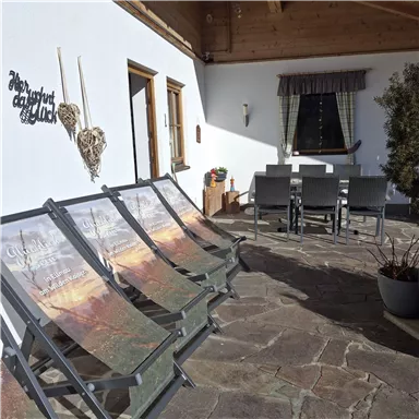 Eine gemütliche Terrasse mit Liegen und einem Esstisch. Im Hintergrund sind Fenster und eine dekorative Wandgestaltung zu sehen.