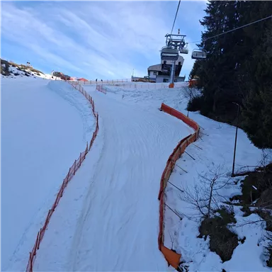 Eine verschneite Skipiste mit einem Skilift im Hintergrund. Der Himmel ist klar und blau, und vor der Piste befinden sich Sicherheitsbarrieren.