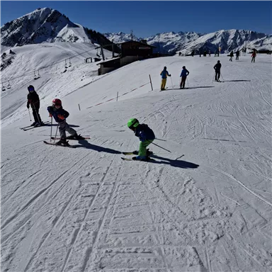 Eine schneebedeckte Landschaft mit skifahrenden Personen. Im Hintergrund sind Berge und ein blauer Himmel sichtbar.