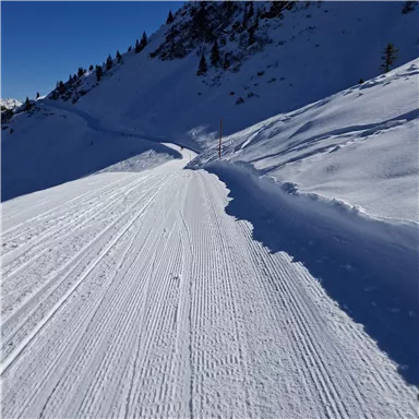 Eine schneebedeckte Skipiste mit frischen Spuren im Schnee. Im Hintergrund sind bergige Landschaften und blauer Himmel zu sehen.