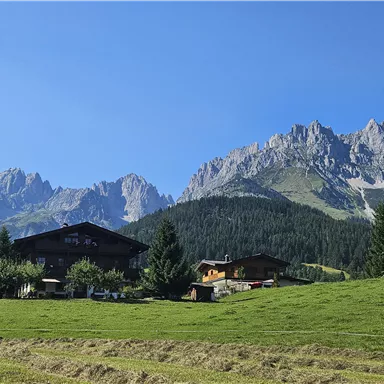 Ein malerisches Bergland mit grünen Wiesen und traditionellen Hütten. Im Hintergrund sind majestätische Berge und ein klarer blauer Himmel zu sehen.