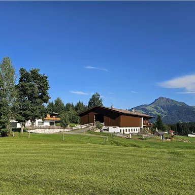 Eine malerische Landschaft mit einem großen Haus und grünen Wiesen. Im Hintergrund erstrecken sich Berge unter einem klaren blauen Himmel.