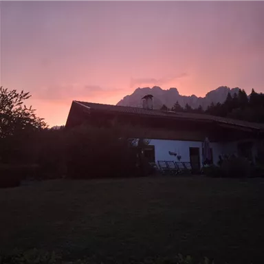 Ein Haus in der Abenddämmerung mit einem bunten Himmel. Im Hintergrund sind majestätische Berge und Bäume zu sehen.