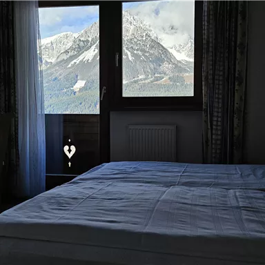 A cozy bedroom with a double bed and a view of the mountains. Daylight streams through the window, showcasing an impressive mountain landscape.