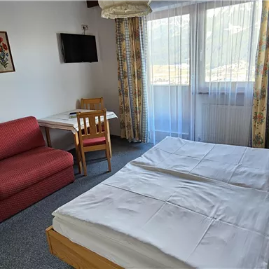 A cozy hotel room with a double bed, a sofa, and a table. Large windows provide plenty of daylight and a pleasant atmosphere.