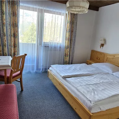 A simple hotel room with two beds and a table. There is a living area with a sofa and a balcony that lets in plenty of daylight.