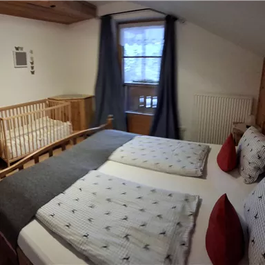 A cozy bedroom with a double bed and a baby cot. The walls are made of wood and the room has large windows with curtains.