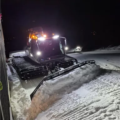 A snow groomer works at night on a snow-covered path. The vehicle's lights shine in the darkness.