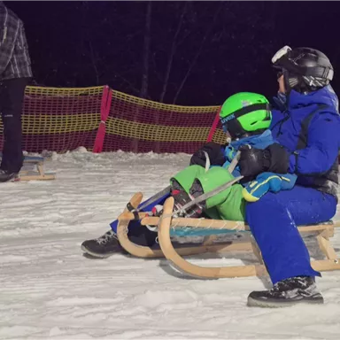 An adult is pulling a sled on a snow track. A child is sitting on the sled, both are wearing winter clothing and helmets.