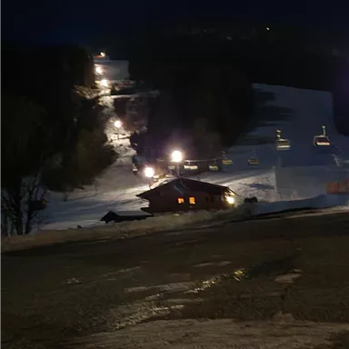 A snowy slope at night with illuminated lifts. In the foreground, a small cabin can be seen.