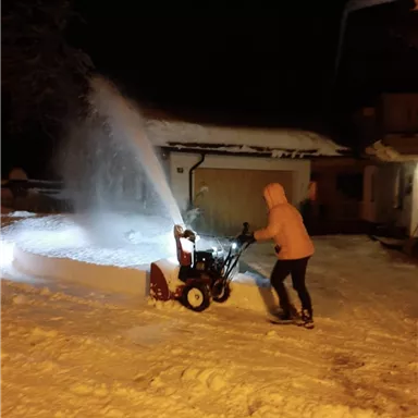 Eine Person bedient eine Schneefräse in einer verschneiten Umgebung bei Nacht. Der Schnee wird in die Luft geblasen, und man sieht ein hell erleuchtetes Haus im Hintergrund.