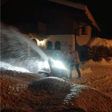Ein Schneepflug räumt Schnee bei Nacht vor einem Haus. Der Schnee wird mit hellem Licht weggeschoben.