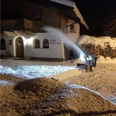 Ein Mann räumt Schnee mit einem Schneeräumer in der Nacht. Im Hintergrund ist ein chaletartiges Haus und ein verschneiter Garten zu sehen.