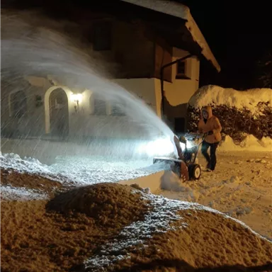 Eine Person räumt mit einer Schneefräse den schneebedeckten Weg vor einem Haus. Es ist Nacht, und das Licht der Maschine erhellt die Umgebung.