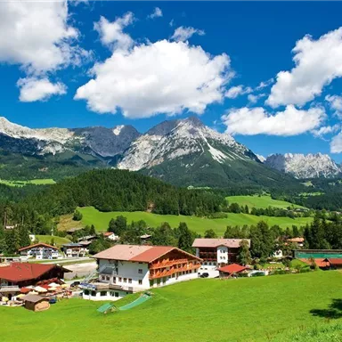 Eine malerische Berglandschaft mit grünen Wiesen und majestätischen Bergen im Hintergrund. Im Vordergrund sind traditionelle alpine Häuser zu sehen, unter einem strahlend blauen Himmel.