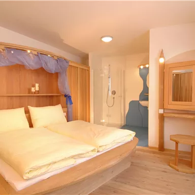 A modern bedroom with a large double bed and wooden surfaces. In the background, there is an adjacent bathroom with a shower.