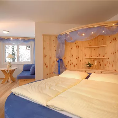 A cozy bedroom with a large bed and light blue bedding. In the background, windows and a small sitting area are visible.