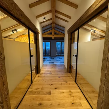 A bright hallway with wooden flooring and wooden frames. On both sides, there are glass doors that lead to separate rooms.