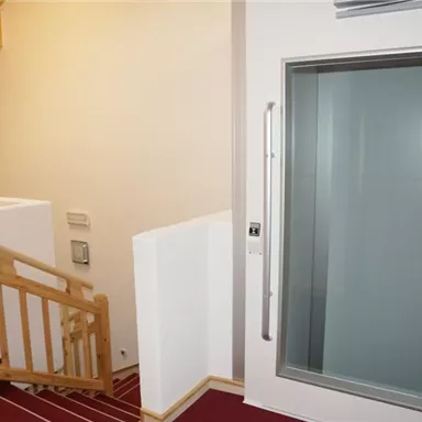 Ein moderner Innenraum mit einer Treppe und einem Glasaufzug. Der Boden ist mit rotem Teppich ausgelegt und die Wände sind hell.