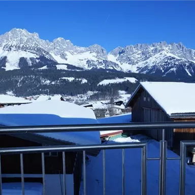 A snow-covered landscape with majestic mountains under a clear blue sky. In the foreground, wooden buildings can be seen that match the wintry scenery.