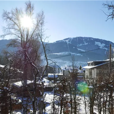 Eine verschneite Landschaft mit einem sonnigen Himmel. Im Hintergrund sind Berge und einige Häuser zu sehen.