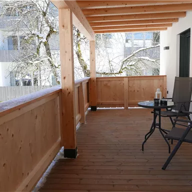 Ein gemütlicher Balkon mit Holzverkleidung, ausgestattet mit einem Tisch und Stühlen. Im Hintergrund sind Bäume und eine helle Wohnanlage zu sehen.