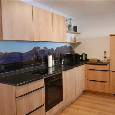 Eine moderne Küche mit Holzschränken und einer dunklen Arbeitsplatte. Im Hintergrund ist ein beruhigendes Bergmotiv zu sehen.