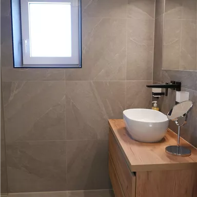 Ein modernes Badezimmer mit grauen Fliesen und einem Holzwaschtisch. Der runde Waschbecken und der große Spiegel sorgen für eine helle, freundliche Atmosphäre.