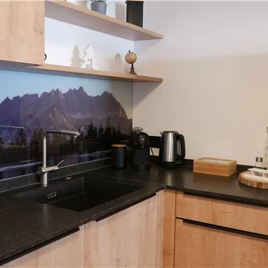 Eine moderne Küche mit Holzregalen und einer dunklen Arbeitsfläche. Im Hintergrund sind Berge und Bäume abgebildet.