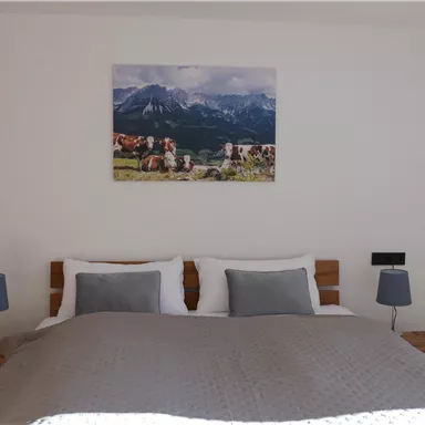 Ein gemütliches Schlafzimmer mit einem großen Bett, zwei Nachttischen und Lampen. An der Wand hängt ein Bild von einer Berglandschaft mit Kühen.