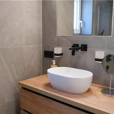 Ein modernes Badezimmer mit grauen Fliesen und einem eleganten Holzwaschtisch. Im Vordergrund steht ein runder Waschbecken und ein Spiegel.