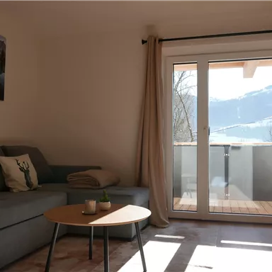Ein gemütlicher Raum mit einem grauen Sofa und einem Tisch. Große Fenster bieten einen Blick auf die Berge und viel Tageslicht.