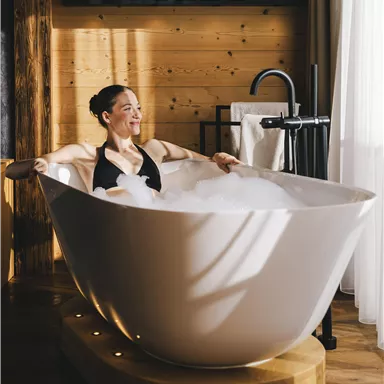 A woman relaxes in a modern bathtub with foam. The room has warm wood and soft lighting.