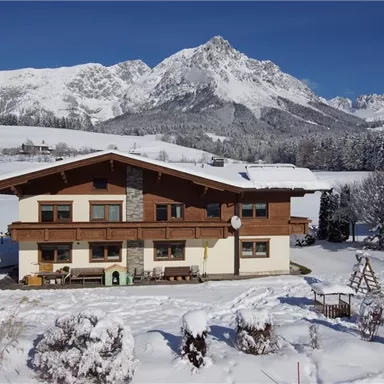 Ein gemütliches Chalet im Schnee, umgeben von schneebedeckten Bergen. Der Himmel ist klar und blau, was eine wunderschöne Winterlandschaft schafft.