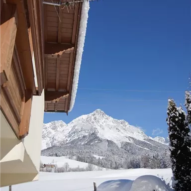 Eine Winterlandschaft mit schneebedeckten Hügeln und Bergen unter einem klaren blauen Himmel. Im Vordergrund ist ein Teil eines Hauses zu sehen.