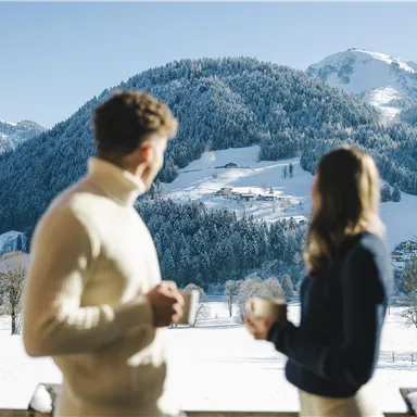 A couple stands on a balcony enjoying hot drinks. In the background, a snowy mountain landscape stretches out under a clear blue sky.