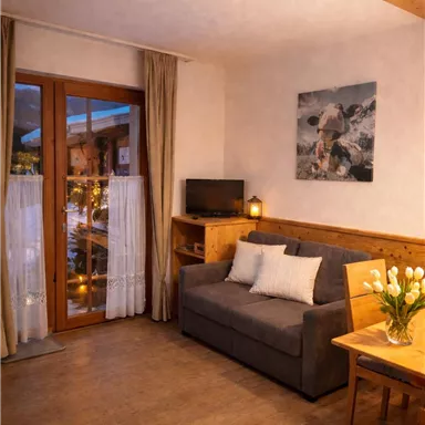 A cozy living room with a gray sofa and a wooden table. Large windows with curtains provide plenty of daylight and a warm atmosphere.