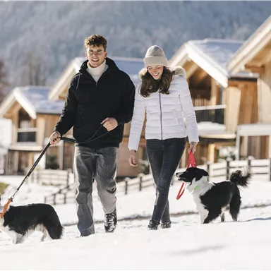 A couple is walking with two dogs through a snowy area. In the background, modern wooden houses and mountains can be seen.