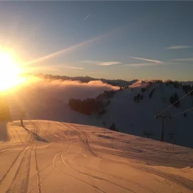 Eine verschneite Berglandschaft bei Sonnenuntergang. Der Himmel ist klar und die Sonne strahlt über den Hügeln.