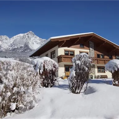Ein wunderschönes, schneebedecktes Haus vor einer malerischen Bergkulisse. Die Umgebung ist ruhig und winterlich.