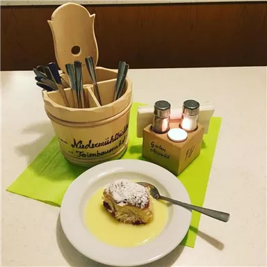 Ein Stück Kuchen mit Puderzucker auf einem Teller, serviert mit einer Vanillesoße. Im Hintergrund befinden sich Besteck und Gewürzbehälter auf einem Tisch.