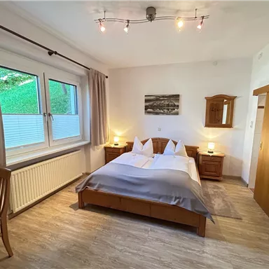 A bright bedroom with a double bed and wooden furniture. The large windows provide plenty of daylight and a cozy atmosphere.