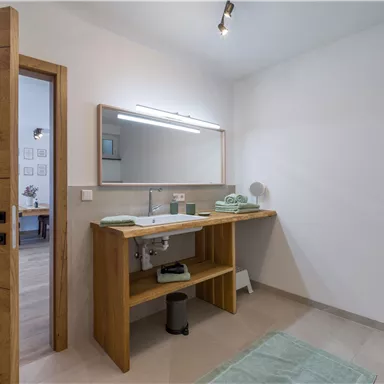 Ein modernes Badezimmer mit einem Holzwaschtisch und einem großen Spiegel. Es gibt grüne Handtücher und einen hellen, minimalistischen Stil.