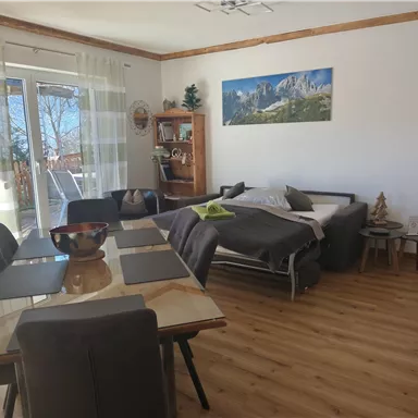 A bright and inviting living room with a dining table and chairs. In the background is a cozy bed and a window with a view of nature.