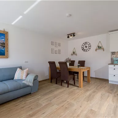 Ein helles Wohnzimmer mit einem blauen Sofa und einem Esstisch. An der Wand hängen Bilder und eine Uhr.