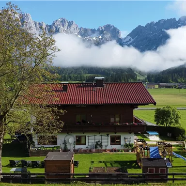 Eine malerische Alm mit einem roten Dach und grüner Wiese. Im Hintergrund erheben sich beeindruckende Berge mit Wolken.