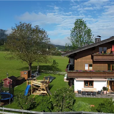 Ein schönes Haus inmitten einer grünen Wiese, umgeben von Bergen. Im Vordergrund gibt es einen Spielplatz und eine Runde Trampolin.