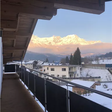 Ein Blick von einem Balkon auf schneebedeckte Berge und ein malerisches Dorf. Die sanften Farben des Sonnenuntergangs verleihen der Szene eine ruhige Atmosphäre.